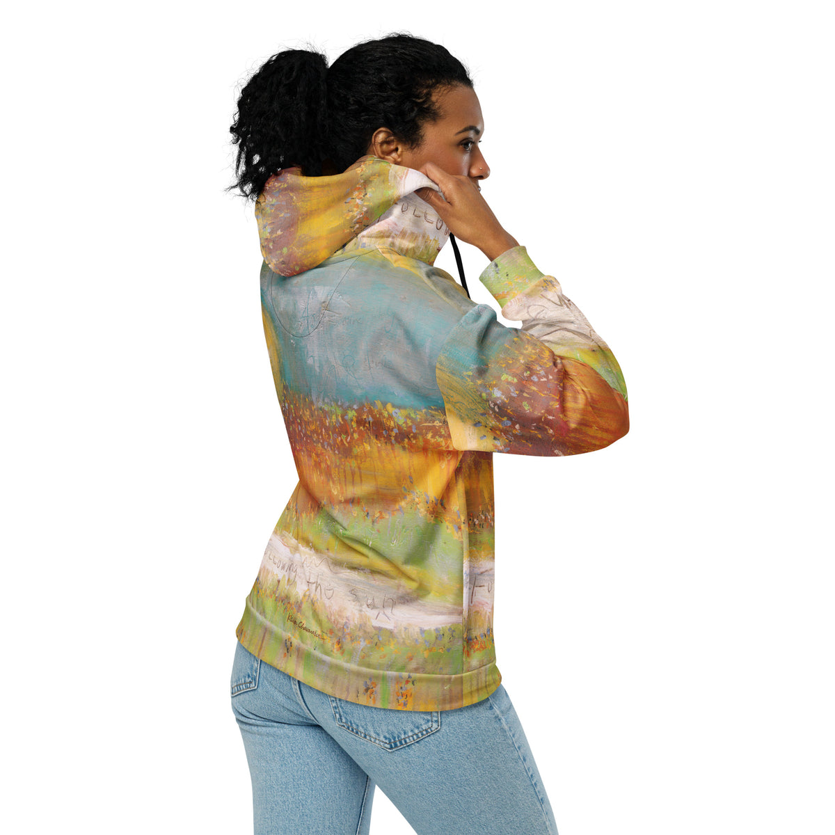 Side back view of a model adjusting the hood of the FOLLOWING THE SUN zip hoodie, painted meadow and handwritten words on the back