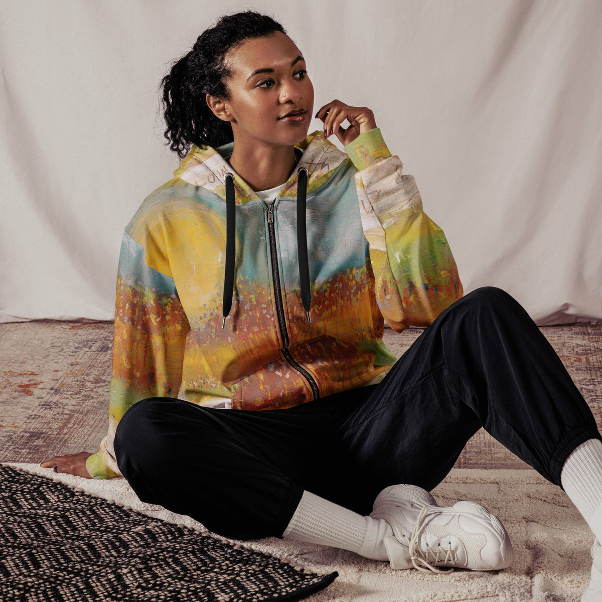 Model seated on a rug in the FOLLOWING THE SUN zip hoodie with black joggers, sun and wildflower print by Klara Chavarría