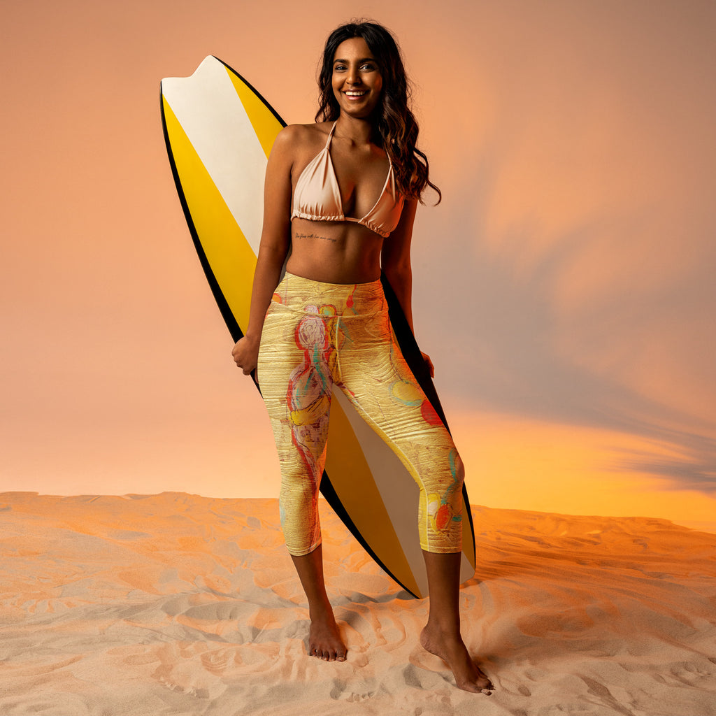 Smiling model on sand holding a yellow and white surfboard, wearing the capris