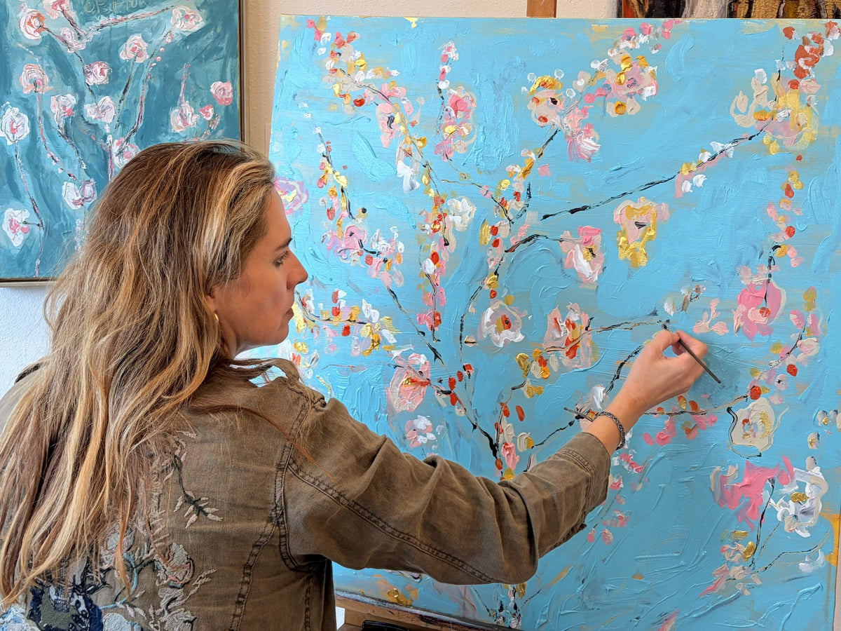 Klara Chavarría painting a blue canvas with pink and yellow blossoms in her studio