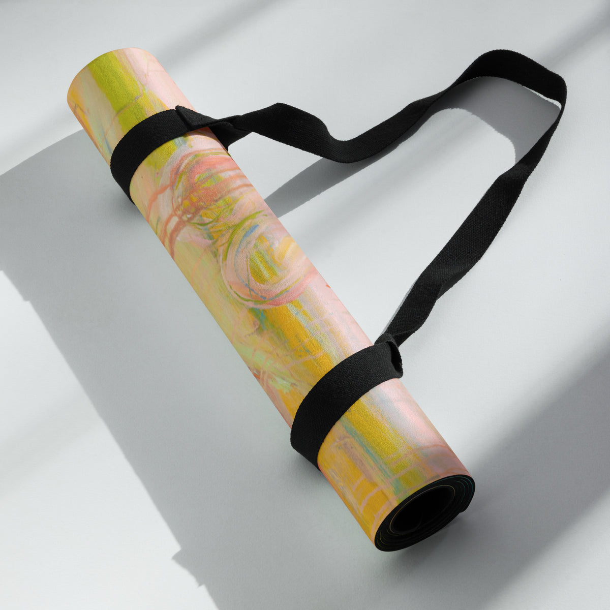 Rolled mat in yellow and pink tones held by a black carrying strap, lying on a white surface